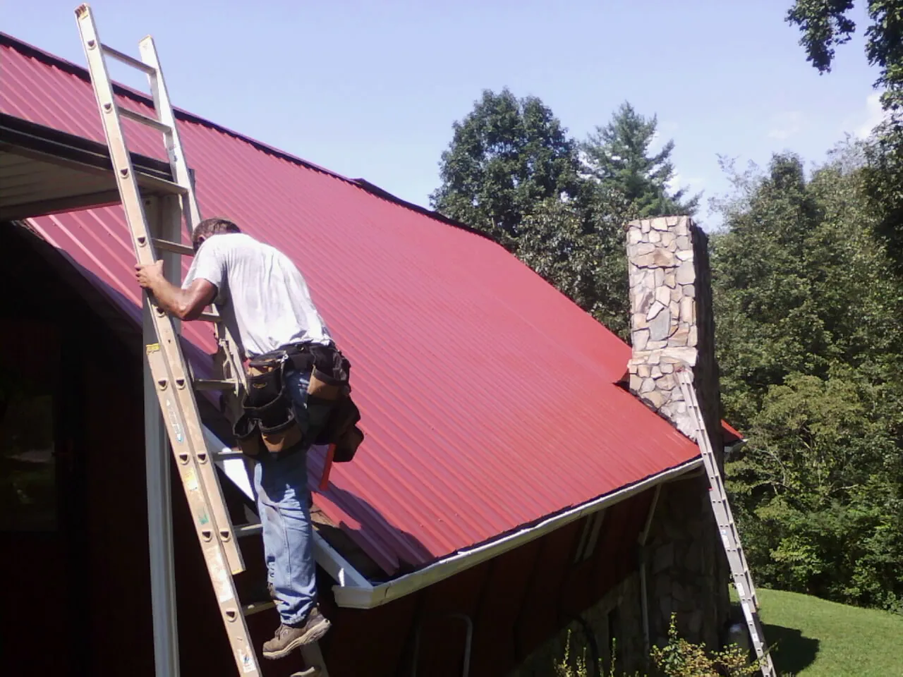 Roofer on a ladder beside a red metal roof and stone chimney — Soffit & Fascia Repair by Roofing Brothers in Norcross
